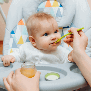 Bébé en repas solide avec une cuillère, aidé par un adulte, dans un cadre familial.